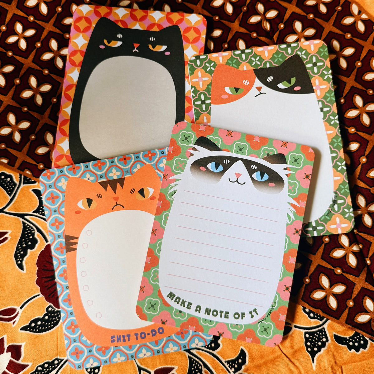 Four cat-themed notepad cards arranged on a patterned orange and brown fabric background. The notepads feature cartoon cat faces, lined writing sections, and motivational phrases like 'MAKE A NOTE OF IT' and 'SHIT TO-DO'.